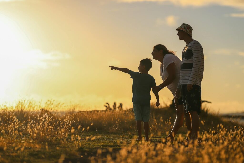 Silhouetted family of three in a sunlit field at sunset; the child points toward something in the distance while the parents watch.