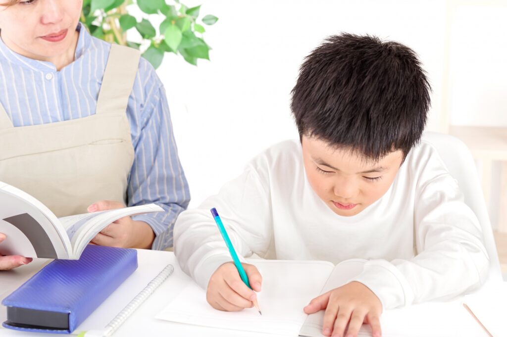 Boy writes in a notebook at a white desk while an adult beside him holds an open book for guidance.